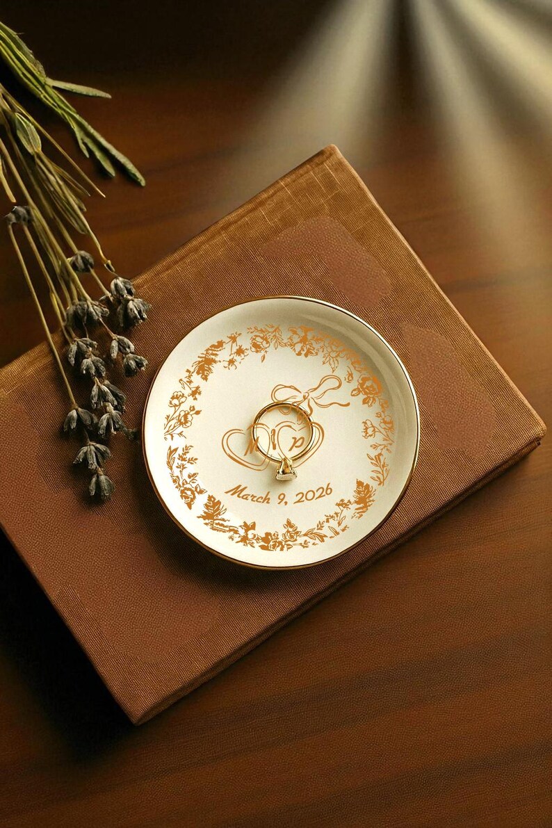 May include: A white ceramic ring dish with gold floral and heart designs, holding a gold ring with a diamond. The dish has a gold rim and the inscription "March 9, 2026". The dish rests on a brown book.