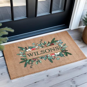 May include: A rectangular doormat made of natural fibers. The mat features the name "WILSONS" in a rectangular frame, surrounded by a festive design of green leaves, red berries, and white flowers, perfect for the holiday season.