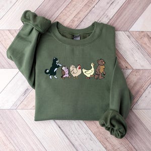 Op de afbeelding: Een olijfgroene sweater met ronde hals en een grafische print van vijf dieren die in een rij lopen. De illustratie toont een zwart-witte kat, een kleine bruine uil, een witte kip, een gele eend en een bruine beer die naar rechts kijken.