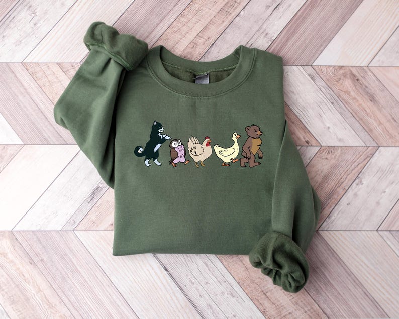 Op de afbeelding: Een olijfgroene sweater met ronde hals en een grafische print van vijf dieren die in een rij lopen. De illustratie toont een zwart-witte kat, een kleine bruine uil, een witte kip, een gele eend en een bruine beer die naar rechts kijken.