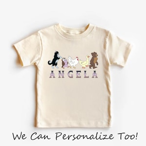 Op de afbeelding: Een cr&egrave;mekleurig T-shirt met korte mouwen en een afbeelding van een zwart-witte kat, een uil, een witte kip, een gele eend en een bruine beer die in een rij lopen. Onder de dieren staat de naam ANGELA gedrukt in paarse letters met een kleurverloop.