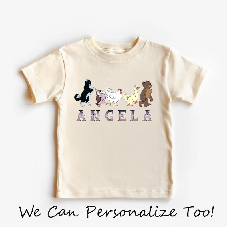 Op de afbeelding: Een cr&egrave;mekleurig T-shirt met korte mouwen en een afbeelding van een zwart-witte kat, een uil, een witte kip, een gele eend en een bruine beer die in een rij lopen. Onder de dieren staat de naam ANGELA gedrukt in paarse letters met een kleurverloop.