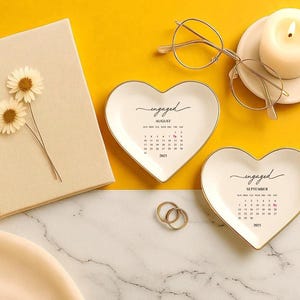 Op de afbeelding: Een stilleven met hartvormige ringschalen met gouden rand, een notitieboek met gedroogde bloemen, een brandende kaars en een bril. De schalen tonen het woord "verloofd" en een kalender.