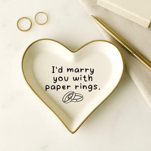May include: A heart-shaped white ceramic trinket dish with a gold rim, featuring the text "I'd marry you with paper rings." Two gold rings are visible. A gold pen and a small box are also present.