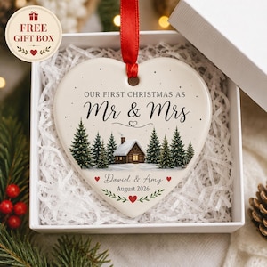 Op de afbeelding: Een hartvormige kersthanger van keramiek met een rood lint, voorzien van een winterse blokhut, dennenbomen en de tekst "OUR FIRST CHRISTMAS AS Mr & Mrs", "David & Amy" en "August 2026" met kleine rode hartjes als decoratie.