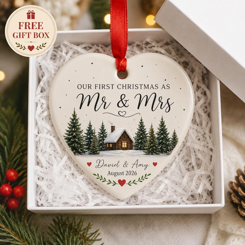 Op de afbeelding: Een hartvormige kersthanger van keramiek met een rood lint, voorzien van een winterse blokhut, dennenbomen en de tekst "OUR FIRST CHRISTMAS AS Mr & Mrs", "David & Amy" en "August 2026" met kleine rode hartjes als decoratie.