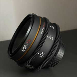 May include: A black camera lens with a gold ring and the text "MIR-1V" in red. The lens has a depth of field scale with markings for 0.6, 0.7, 0.8, and 3. The lens is made in Ukraine.