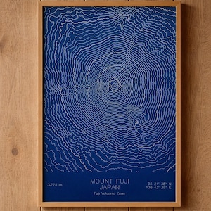 May include: A framed print featuring a detailed topographic map of Mount Fuji, Japan. The map is rendered in white lines against a deep blue background. The frame is made of light-colored wood. The text on the print includes the mountain's elevation, 3,775 m.