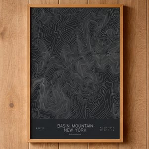 May include: A framed wall art print featuring a black background with white topographic contour lines of Basin Mountain in New York. The text reads Basin Mountain New York, 4,827 ft, and the coordinates 44 07 16 N 73 53 11 W in white.