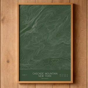 May include: A framed topographic map print of Cascade Mountain in New York, featuring white contour lines on a dark green background. The text at the bottom reads Cascade Mountain, New York, 4,098 ft, and the coordinates 44 13 07 N 73 51 38 W.