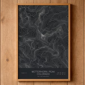 May include: A rectangular framed wall art print featuring a black background with white topographic contour lines of Wetterhorn Peak in the San Juan Mountains, Colorado. The text reads 14,021 ft, Wetterhorn Peak, Colorado, San Juan Mountains, 38 03 39 N, 107 30 39 W.