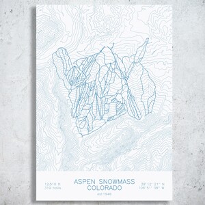 May include: A white print featuring blue topographic contour lines of the Aspen Snowmass ski area in Colorado. The text reads ASPEN SNOWMASS COLORADO, est 1946, 12,510 ft, 319 trails, and the coordinates 39 12 21 N 106 51 38 W in blue font.