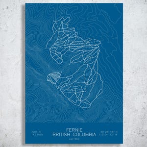 May include: A blue rectangular print featuring white topographic contour lines of a mountain landscape. The text reads FERNIE BRITISH COLUMBIA, 7001 ft, 142 trails, and est 1962, with coordinates 49 28 48 N 115 04 12 W printed at the bottom.