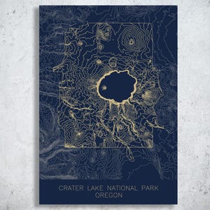 May include: A rectangular print featuring a dark navy blue background with gold topographic contour lines mapping Crater Lake National Park in Oregon. The text CRATER LAKE NATIONAL PARK OREGON is printed in a clean, sans-serif font at the bottom.