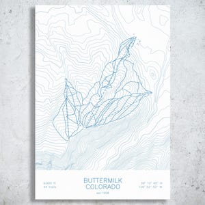 May include: A white print featuring blue topographic contour lines of Buttermilk, Colorado. The text reads: BUTTERMILK COLORADO, est 1958, 9,900 ft, 44 trails, and coordinates 39 10 45 N 106 52 50 W. The design displays ski trail paths.