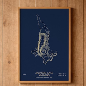 May include: A framed wall art print featuring a dark navy blue background with gold-colored topographic contour lines of Jackson Lake, Wyoming. The text reads Jackson Lake, Wyoming, Grand Teton National Park, 457 ft, and coordinates.