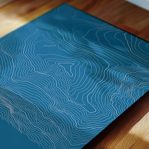 May include: A framed print featuring a topographic map design. The artwork showcases white contour lines against a teal background. The print is displayed on a wooden surface, creating a contrast in textures and colors.