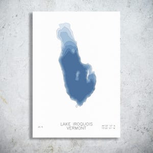 May include: A white wall art print featuring a blue bathymetric map of Lake Iroquois in Vermont. The map displays varying shades of blue to indicate depth, with the text 'LAKE IROQUOIS VERMONT', '25 ft', and coordinates '44°22' 12" N 73°05' 01" W'.