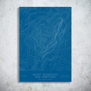 May include: A blue topographic map print of Mount Monadnock in New Hampshire. White contour lines detail the mountain's elevation. The print includes the mountain's name, coordinates, and elevation of 3,165 ft.