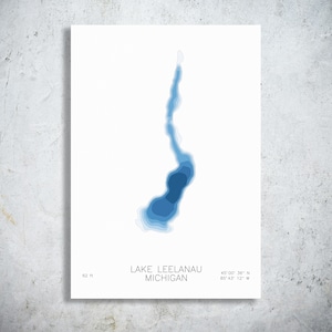 May include: A white rectangular print featuring a blue bathymetric map of Lake Leelanau, Michigan. The text reads Lake Leelanau Michigan, 62 ft, and the coordinates 45 00 36 N 85 43 12 W. The lake shape uses varying shades of blue to show depth.
