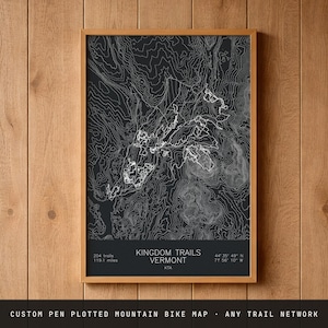 May include: A black poster featuring white topographic contour lines of the Kingdom Trails in Vermont. The text reads Kingdom Trails Vermont, 204 trails, 119.1 miles, and coordinates 44 35' 49" N 71 56' 10" W. The map is in a wooden frame.