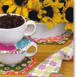 May include: Image of a white coffee cup filled with coffee beans, resting on a floral coaster. The image also shows a folded patterned napkin and several hexagon-shaped hot pads. The text at the top reads "Amazingly Simple Coasters & Hot Pads."