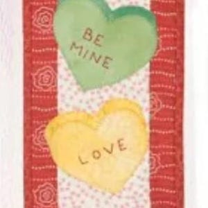 May include: A Valentine's Day-themed fabric banner featuring four heart-shaped candies with the words "HUG", "BE MINE", "LOVE", and "KISS". The banner has a red floral patterned border and a white background with small pink dots.