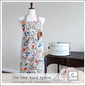 May include: A white apron with a floral print featuring red, pink, yellow, and blue flowers. The apron has a large pocket on the front and a wooden spoon tucked into the pocket. The apron is hanging on a white mannequin. The text "The One Yard Apron" and "Fresh designs from a Wyoming farm." is at the bottom of the image. The image also includes a white cake carrier with the word "PATISSERIE" on it.