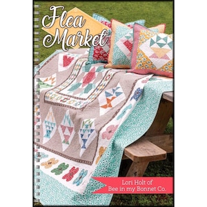 May include: A book titled "Flea Market" by Lori Holt of Bee in my Bonnet Co. The book cover features a quilt with a variety of colorful patterns and a picnic table with pillows.