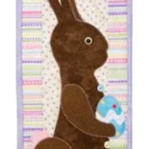 May include: A decorative Easter-themed wall hanging featuring a brown bunny holding a blue egg with white accents. The bunny has pink inner ears and is set against a background of pastel stripes and polka dots, framed by a lavender border.