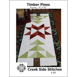 May include: A table runner featuring a Christmas tree design with green and white fabric. The runner is 16 inches by 40 inches and has a red star in the center. The text "Timber Pines" and "Creek Side Stitches #365" is visible.