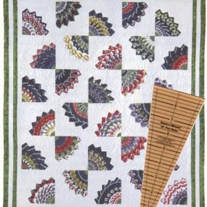 May include: A handmade quilt with a white background and a repeating fan pattern in various colors, including red, green, and purple. The quilt has a green border and a wooden fan ruler is placed on the side.