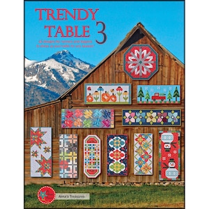 Puede incluir: Una portada de libro con el título "Trendy Table 3" y un fondo de granero rústico. La portada presenta 15 diseños de colchas coloridos para caminos de mesa y cubiertas.
