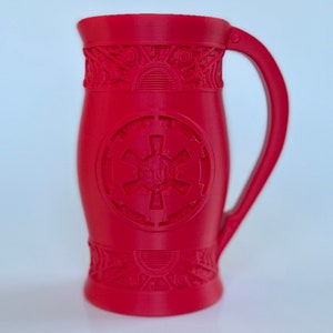 May include: A red ceramic mug with a raised design of the Star Wars Imperial symbol. The mug has a handle and intricate details around the top and bottom.