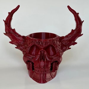 May include: A red skull-shaped planter with two large horns. The planter is made of a textured material and has a detailed design.