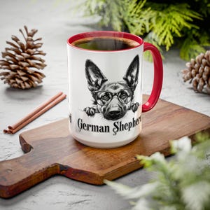 Pode incluir: Caneca de cerâmica branca com borda e alça vermelhas. A caneca apresenta uma ilustração em preto e branco de um pastor alemão usando óculos e espiando por cima da borda da caneca. O texto "German Shepherd" está impresso na caneca.