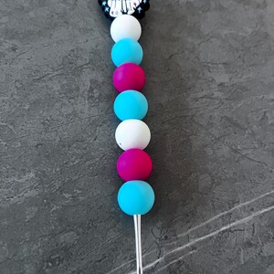 May include: A decorative skewer with a black and white charm featuring a ghost wearing a Santa hat, and the text "BEAUS BUT IT'S CHRISTMAS". The skewer has alternating white, blue, and pink spheres.