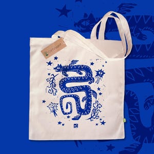 Könnte beinhalten: Eine cremefarbene Stofftasche mit einem blauen Grafikdruck eines geflügelten, schlangenartigen Wesens, umgeben von Sternen und abstrakten Formen. Ein braunes Pappetikett mit der Aufschrift Earth-ware ist am oberen Henkel befestigt.
