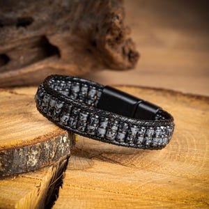 May include: A bracelet featuring two parallel rows of small, cylindrical, grey and black speckled stone beads. The beads are framed by braided black leather cords on the top and bottom edges, secured with a smooth, rectangular black metal clasp.