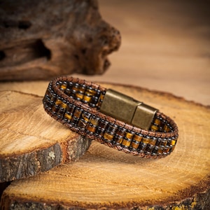 Könnte beinhalten: Ein Armband mit Reihen kleiner, zylindrischer Tigeraugen-Perlen in Braun- und Goldtönen, die mit einer dunklen Kordel verflochten sind. Das Schmuckstück wird mit einem rechteckigen, magnetischen Metallverschluss in Antik-Messing-Optik geschlossen.