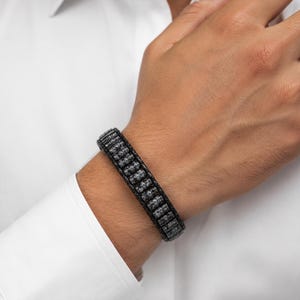 Könnte beinhalten: Ein schwarzes Armband mit einer mittleren Reihe aus kleinen, zylindrischen, metallisch glänzenden Perlen, die von zwei geflochtenen schwarzen Bändern flankiert werden. Das Design zeigt ein Muster aus dunklen, texturierten Elementen.