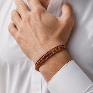 Op de afbeelding: Een armband met een enkele rij doorschijnende, ronde, barnsteenkleurige kralen die tussen twee parallelle bruine koorden zijn bevestigd. De kralen hebben een gladde, gepolijste afwerking en een warme, goudbruine tint in een lineair ontwerp.