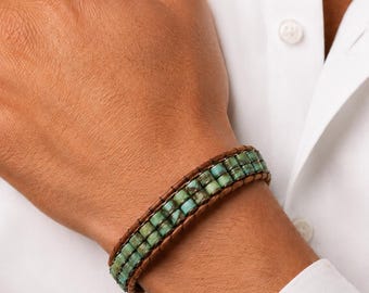 Op de afbeelding: Een armband met meerdere strengen, bestaande uit twee rijen kleine, vierkante, turquoise kralen met donkere aders, vastgezet tussen twee parallelle strengen donkerbruin leer die met een dunne bruine draad aan elkaar zijn genaaid.