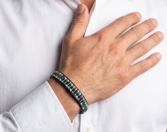 Op de afbeelding: Een wikkelarmband met meerdere strengen, voorzien van rijen kleine, vierkante turquoise en donker metallic kralen. De kralen zijn in een herhalend patroon gerangschikt en vastgezet met een donker koord, wat een gestructureerde band vormt.