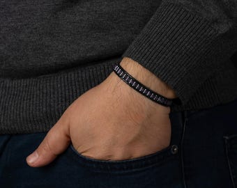 May include: A slim black bracelet featuring a series of small, rectangular purple stones or beads set in a row. The bracelet has a dark, woven-style band and is worn around the wrist, contrasting against a dark grey sweater and dark blue jeans.