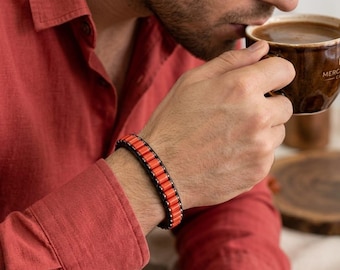 May include: A bracelet featuring a row of bright orange cylindrical beads secured between two parallel black leather cords, held together by small metallic rivets along the edges, creating a structured and layered appearance on the wrist.