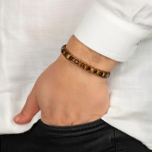 May include: A bracelet featuring a single row of rectangular tiger eye beads strung on a dark cord. The beads display natural golden-brown chatoyant bands and are arranged in a continuous line, secured with a simple knot closure mechanism.