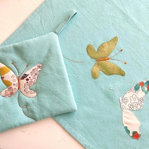 Peut inclure: Un gant de cuisine en tissu bleu clair et un torchon assorti, tous deux ornés de motifs de papillons en appliqué. Les papillons sont confectionnés à partir de tissus à motifs variés dans des tons de vert, blanc, orange et marron, avec des détails brodés.