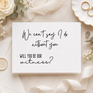 May include: A white rectangular card with black text that reads: We can't say I do without you WILL YOU BE OUR witness? The card is surrounded by white roses, baby's breath, a wax seal, and two gold rings on a white scalloped dish.