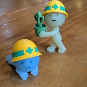 Puede incluir: Dos peque&ntilde;as figuras, una azul y otra verde p&aacute;lido, ambas con cascos amarillos con rayas verdes y una cruz blanca. La figura verde sostiene un peque&ntilde;o cactus verde en maceta. La figura azul est&aacute; en el suelo.